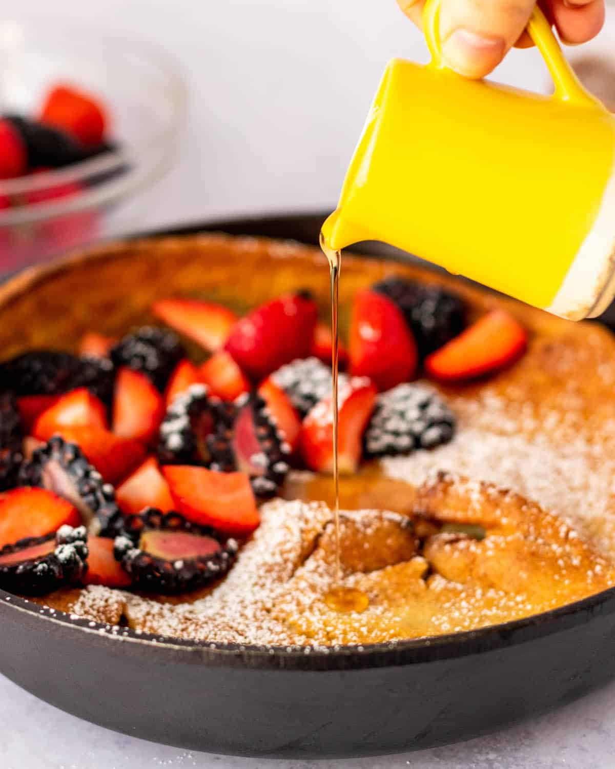 pouring maple syrup over gluten free dutch baby with berries in skillet