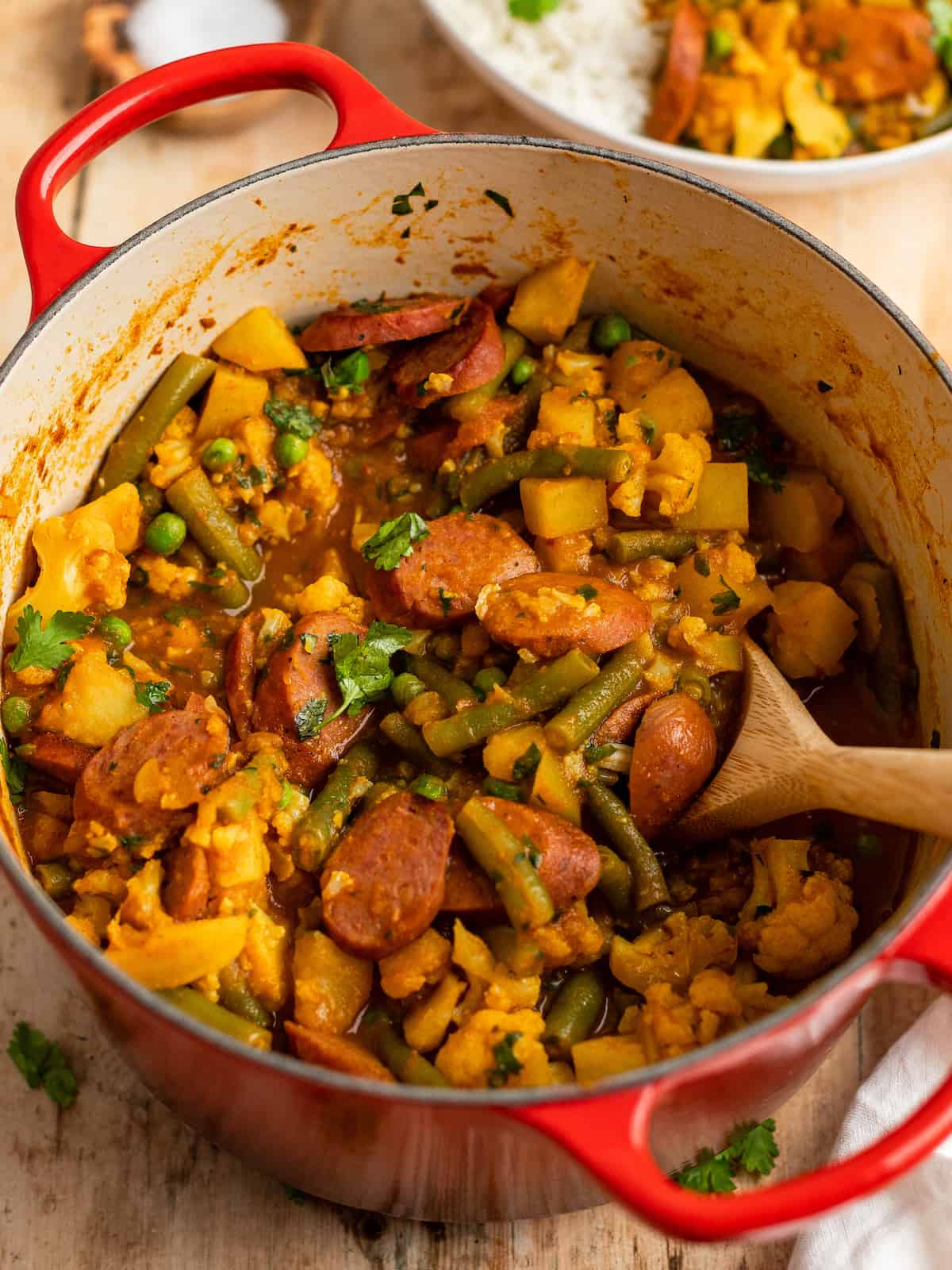 sausage curry in a red dutch oven with a wooden spoon