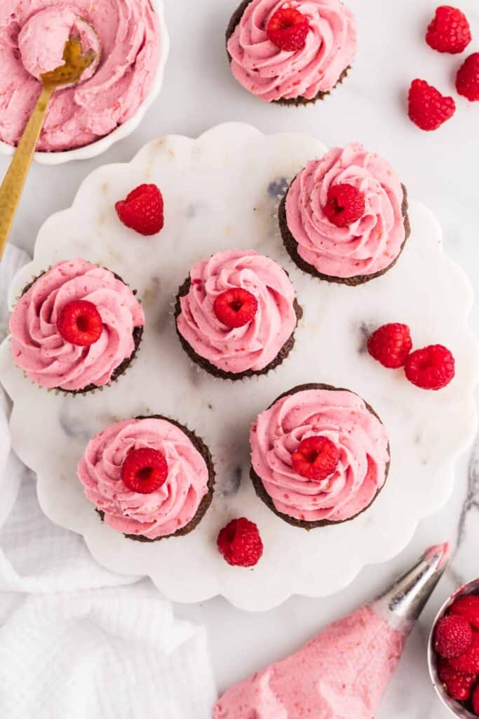 Overhead view of six chocolate cupcakes topped with piped pink raspberry cheesecake frosting and fresh raspberries on a marble cake stand