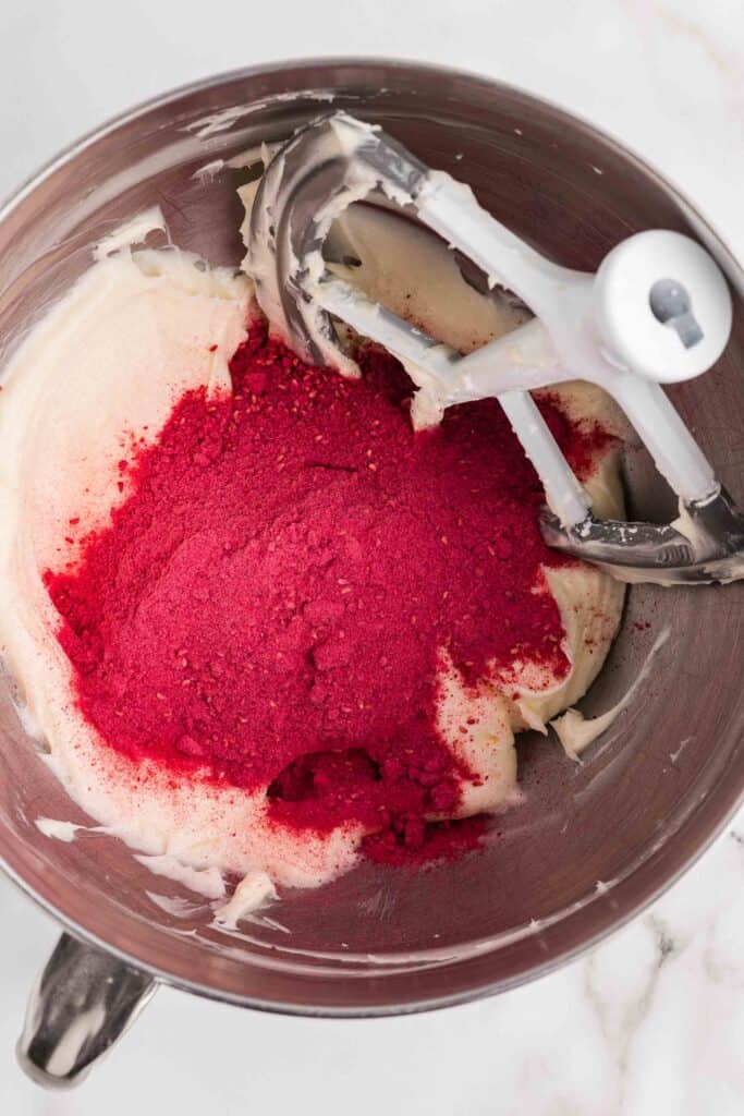 Freeze-dried raspberry powder being added to the cream cheese mixture in a stand mixer bowl