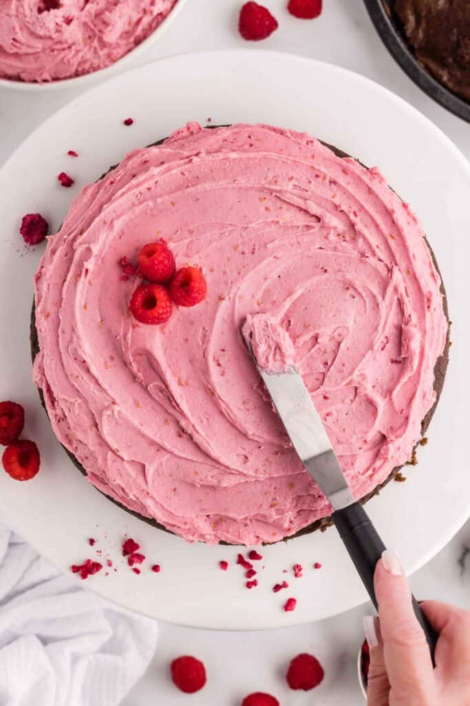 Overhead view of a chocolate cake being frosted with pink raspberry cheesecake filling and garnished with fresh raspberries