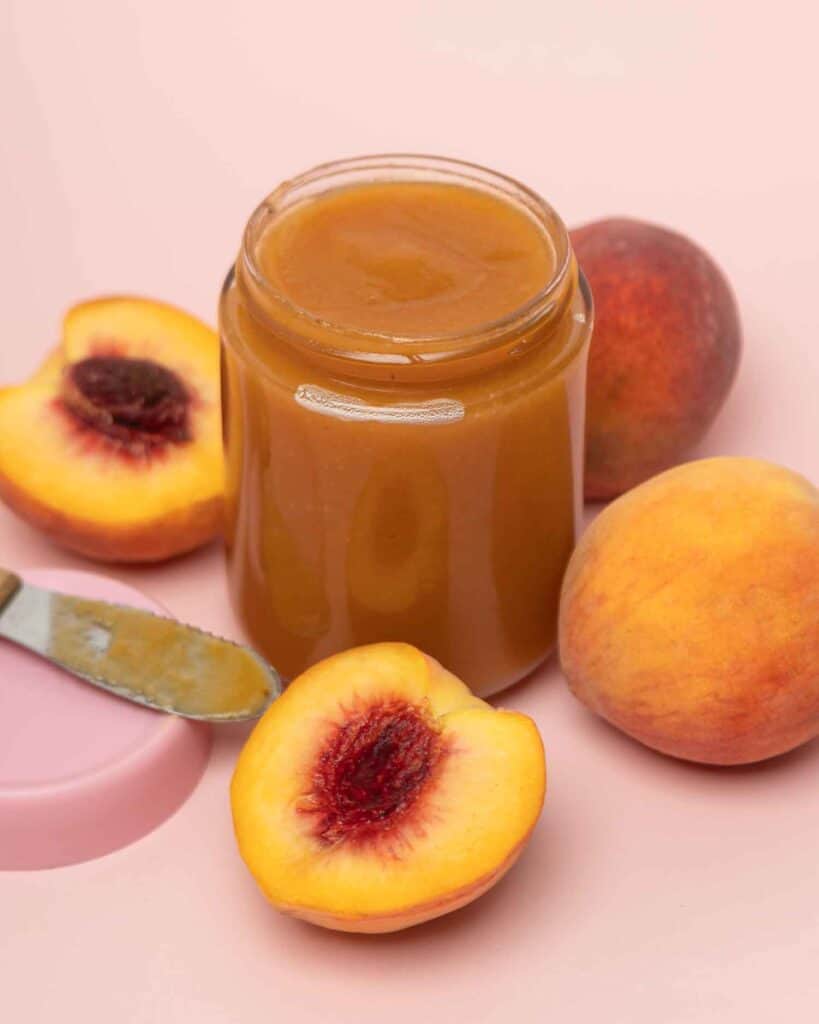 Jar of crockpot peach butter with fresh halved peaches on a pink background