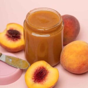 Glass jar of homemade crockpot peach butter surrounded by fresh ripe peaches