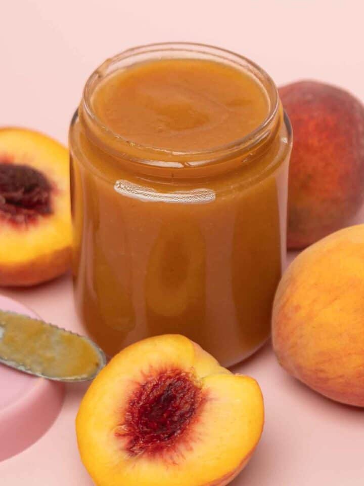 Glass jar of homemade crockpot peach butter surrounded by fresh ripe peaches