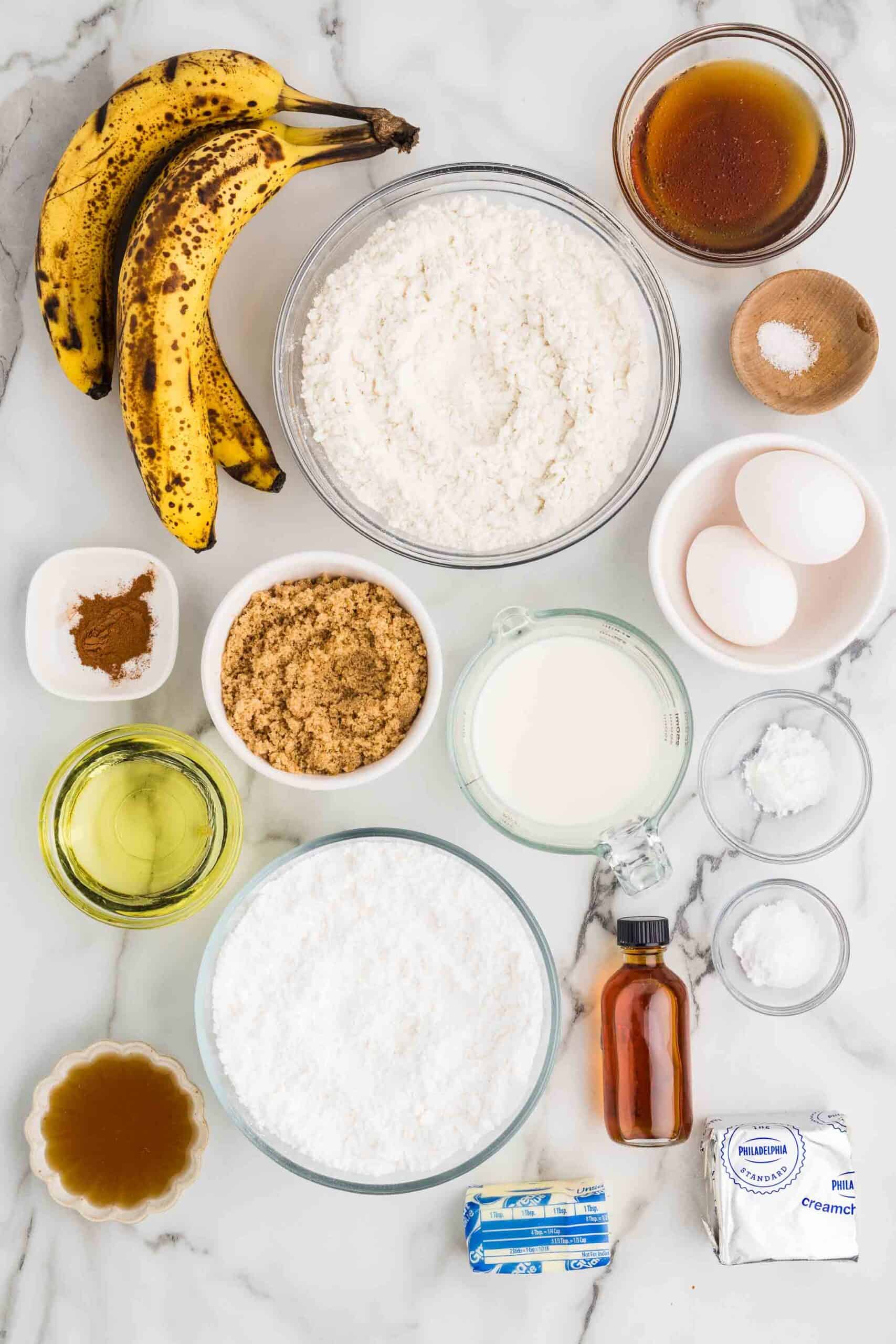 Ingredients for gluten-free banana cake on a marble counter: ripe bananas, gluten-free flour, brown sugar, maple syrup, milk, eggs, oil, cream cheese, butter, powdered sugar, and caramel
