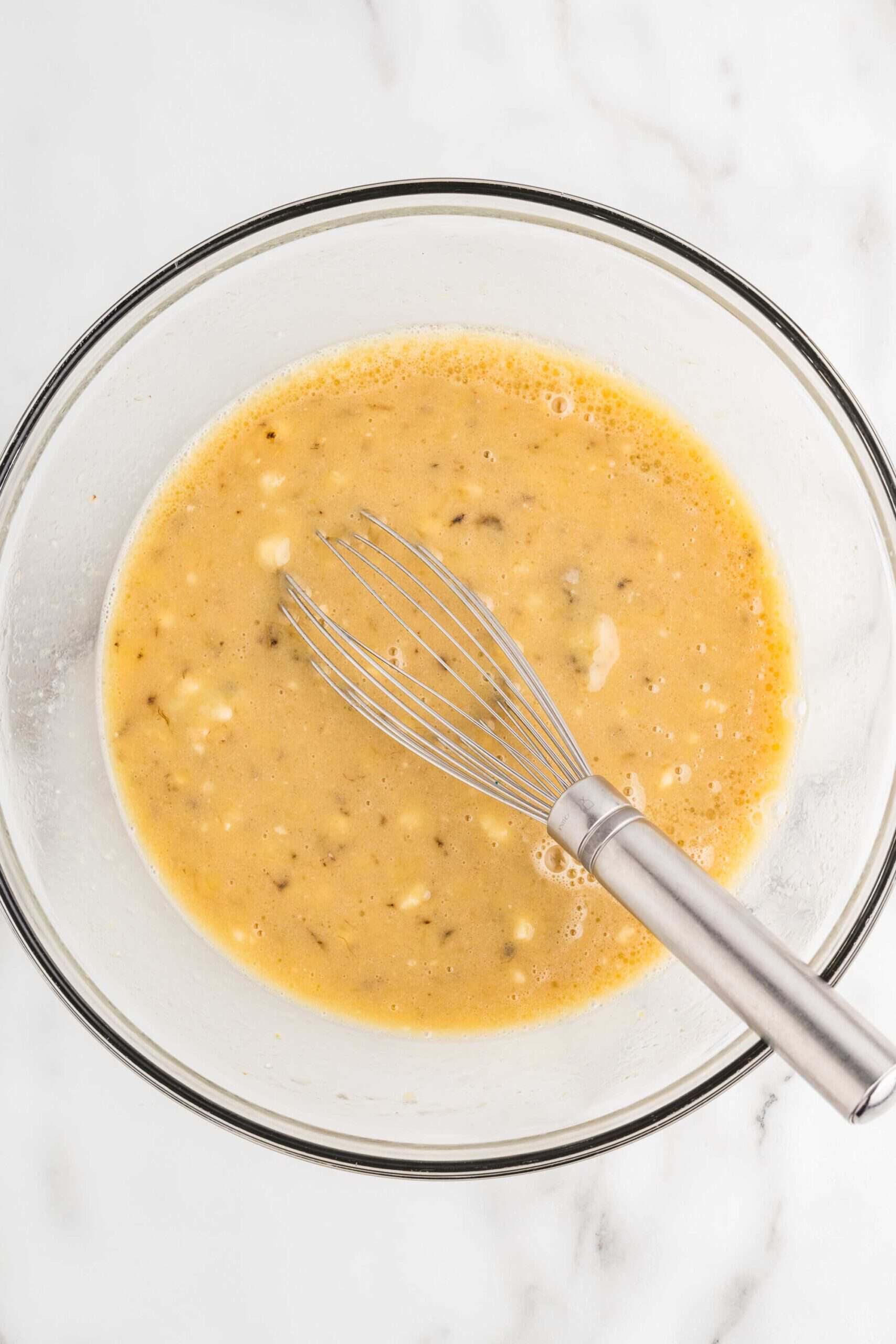 Mashed banana, oil, sugar, maple syrup, milk, and eggs whisked together in a glass bowl