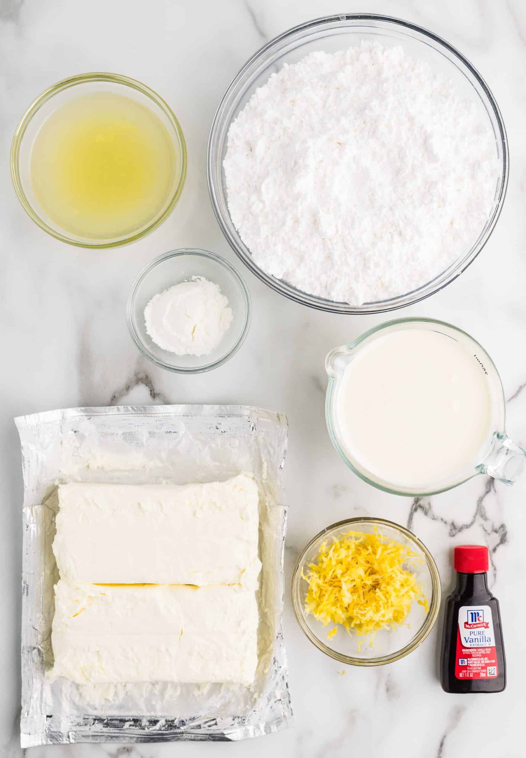 Ingredients for lemon cheesecake filling on a marble counter: cream cheese, heavy cream, powdered sugar, cornstarch, lemon juice, lemon zest, and vanilla extract