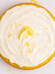 Overhead view of a round lemon cake topped with swirls of no bake lemon cheesecake filling, lemon zest, and a lemon slice