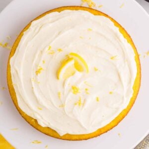 Overhead view of a round lemon cake topped with swirls of no bake lemon cheesecake filling, lemon zest, and a lemon slice