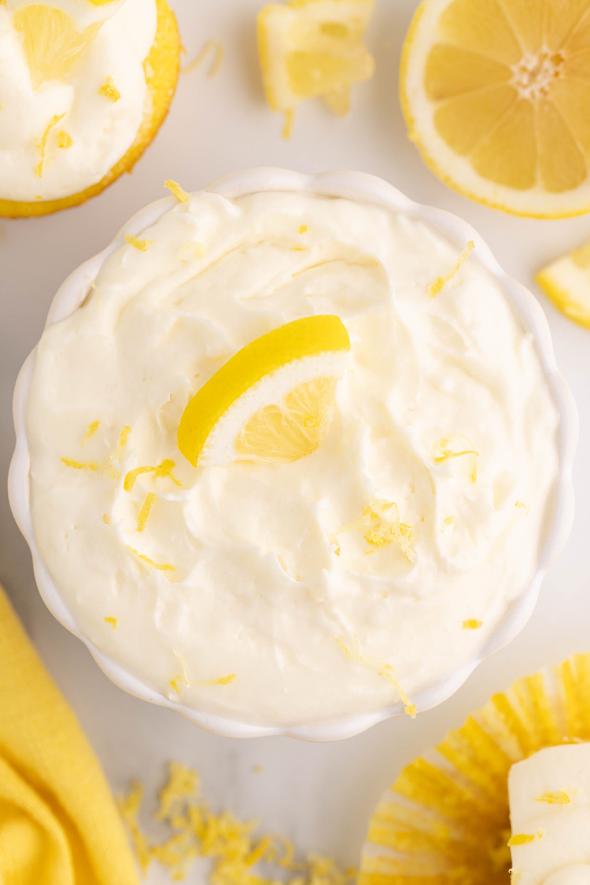 A bowl of fluffy lemon cheesecake filling topped with lemon zest and a lemon slice, surrounded by lemons